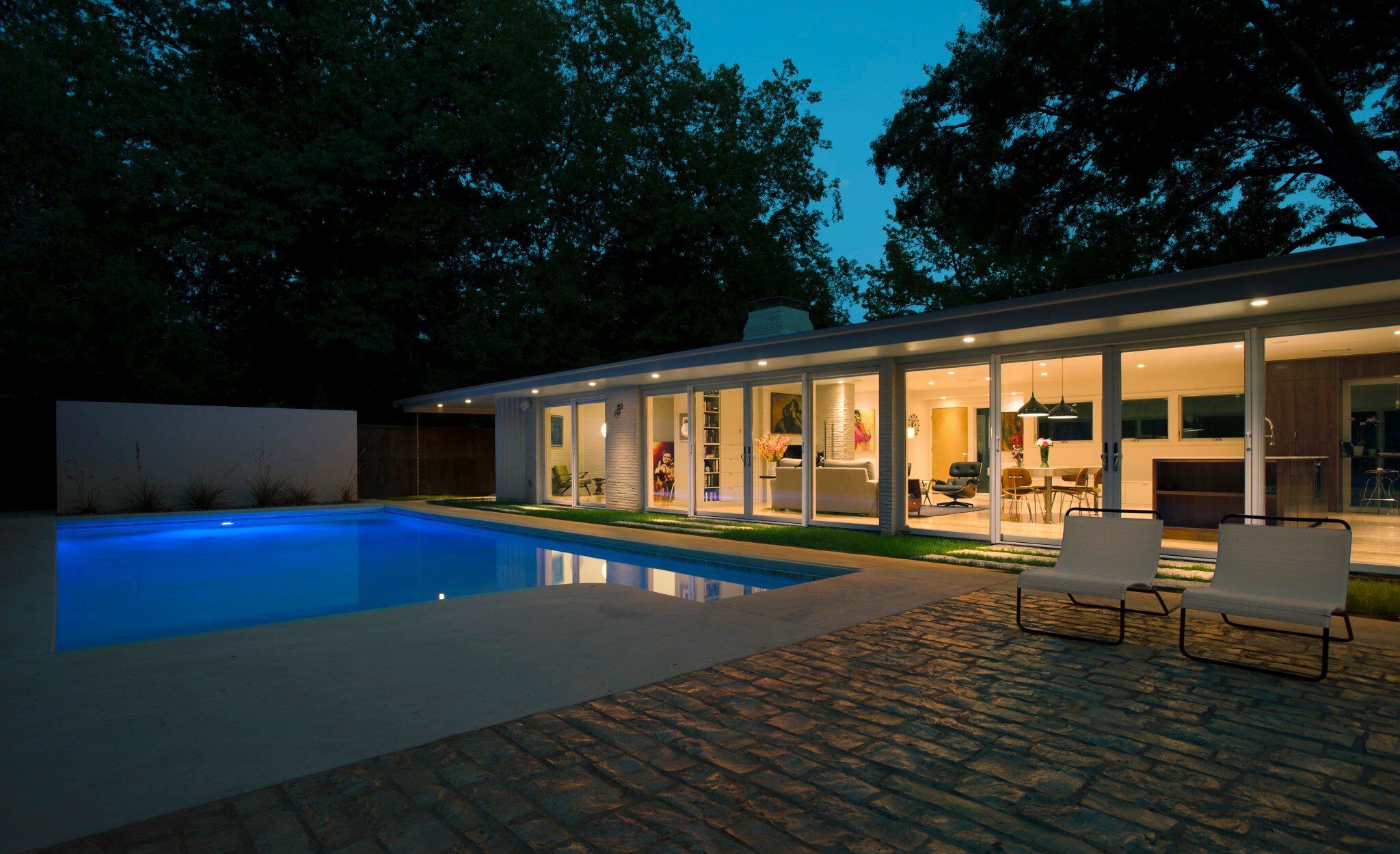 Mid-century modern home exterior. View of back yard and custom pool at night, with view into living spaces through walls of windows. Home renovation by Brent Swift and SwiftCo in Norman, Oklahoma. Purpose is to show our quality of work.